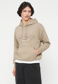 Χακί φούτερ με μπροστινή τσέπη και τόνους σχεδίου λογότυπου. Διαθέτει κουκούλα με κορδόνι και ribbed μανσέτες. Συνδυασμένο με σκούρα παντελόνια.