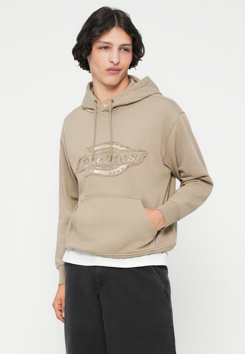 Khaki hoodie with a front pocket and tonal logo design. Features a drawstring hood and ribbed cuffs. Paired with dark pants.