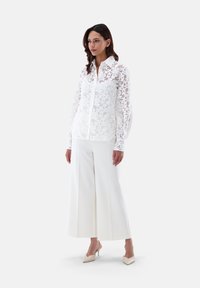 White floral lace blouse with button front, long sleeves, and a collared neck, paired with wide white trousers and nude heels.