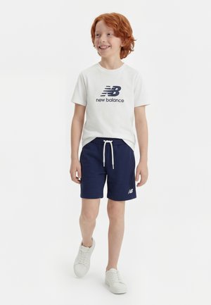 Ragazzo dai capelli rossi che cammina, indossa una maglietta bianca New Balance, pantaloncini blu navy e sneakers bianche, sorride e guarda di lato.