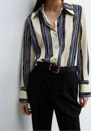 Vrouw die een blouse met lange mouwen draagt, met verticale zwarte en blauwe strepen, ingestopt in een zwarte broek met hoge taille en een zwarte riem.