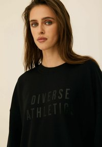 Czarny sweter z okrągłym dekoltem, z matowym czarnym nadrukiem "Diverse Athletics". Miękka tekstura i luźny krój.
