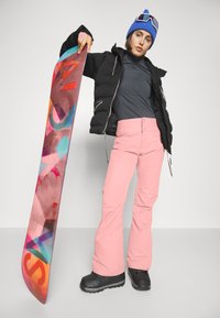 Schwarze Pufferjacke, dunkelgraues Oberteil, rosa Schneehose und schwarze Schneestiefel; hält ein hellbuntes Snowboard mit abstrakten Mustern.