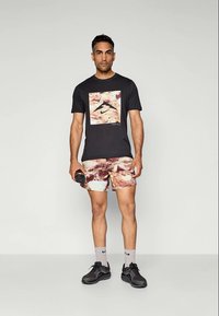 Mann iført svart grafisk t-skjorte og mønstrede shorts holder en vannflaske, stående mot en ensfarget lys bakgrunn.