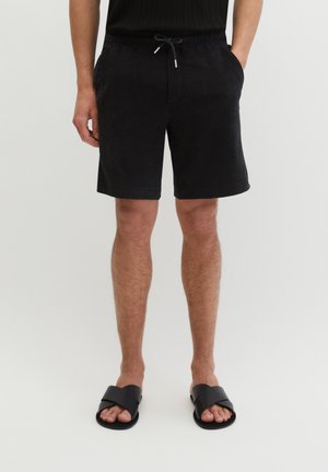 Mężczyzna ubrany w czarne szorty z sznurkiem i czarne sandały z krzyżującymi się paskami, stojący z rękami w kieszeniach na jednolitym tle.