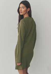 Robe côtelée vert olive à manches longues, présentant une coupe décontractée, des coutures latérales et un ourlet légèrement arrondi. La texture est douce et extensible.