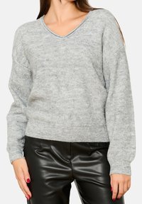 Grauer Strickpullover mit V-Ausschnitt, gerippten Bündchen und Saum. Weiche Textur mit dezentem Schimmer. Kombiniert mit schwarzen Kunstlederhosen.