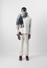 Vit zip-up jacka med hög krage, krämfärgad och grönrutig halsduk, ljusa byxor och burgundy träningsskor. Reflexsolglasögon och fickdetalj.