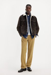 Jachetă din corduroy maro cu guler din blană, suprapusă peste o cămașă din denim, asociată cu pantaloni ușor khaki și încălțăminte neagră.