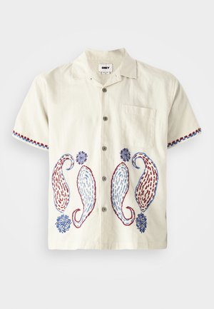 Camicia a maniche corte, di colore beige chiaro, realizzata in cotone con ricami paisley in blu e rosso. Caratterizzata da una tasca sul petto e chiusura con bottoni.