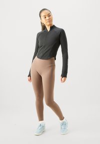 Ung kvinna klädd i en svart zip-jacka, bruna leggings, vita strumpor och ljusblå sneakers som står framför en enfärgad bakgrund.