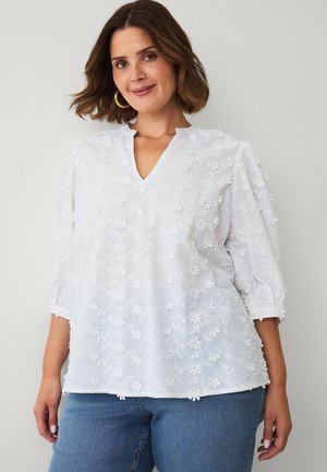Kvinde med skulderlangt brunt hår iført hvid bluse med blomstertekstur og 3/4-ærmer samt blå jeans, stående mod en ensfarvet baggrund.