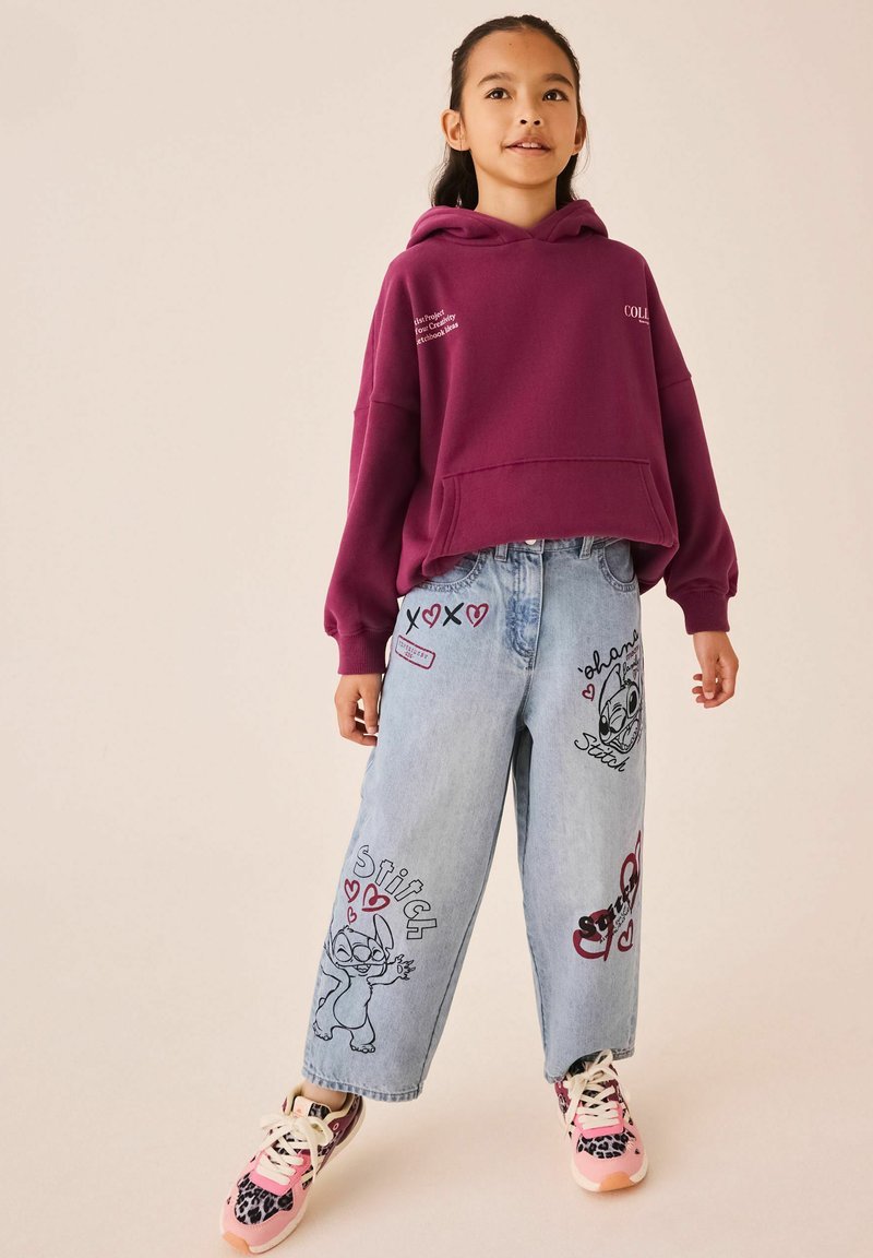 Jeune fille portant un sweat à capuche bordeaux et un jean bleu clair avec des imprimés du dessin animé Stitch, ainsi que des baskets roses, debout devant un fond uni.