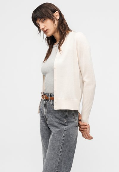 Γυναίκα με σκούρα μαλλιά που φοράει κρεμ ζακέτα με κουμπιά, γκρι εφαρμοστό τοπ, ψηλόμεσα γκρι τζιν και καφέ ζώνη, σε λευκό φόντο.