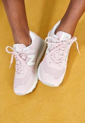 Stopy w jasnoróżowych sneakersach New Balance z białymi akcentami na musztardowożółtej teksturowanej podłodze.
