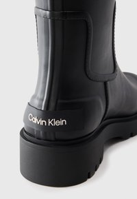 Botte de cheville noire à semelle épaisse avec un talon strié, marquée "Calvin Klein" en blanc sur le petit onglet noir au talon, finition mate lisse.