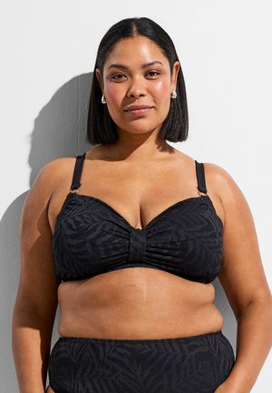 Žena s kratkom crnom kosom nosi crni kupaćI kostim s uzorkom, stojeći nasuprot bijelog zida s bacenom sjenom iza sebe.