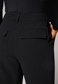 Pantalones negros ajustados con una textura suave, frente plano y bolsillos traseros con cierres de solapa. Marca sutil en el bolsillo.