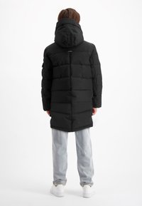 Zwarte gewatteerde jas met capuchon en horizontaal gestikt patroon, gecombineerd met lichtblauwe jeans en witte schoenen.