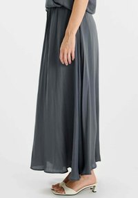 Grijze, tot op de grond vallende rok gemaakt van lichtgewicht stof, met een gladde textuur en een elastische tailleband. Draagt bij lichte, open sandalen.