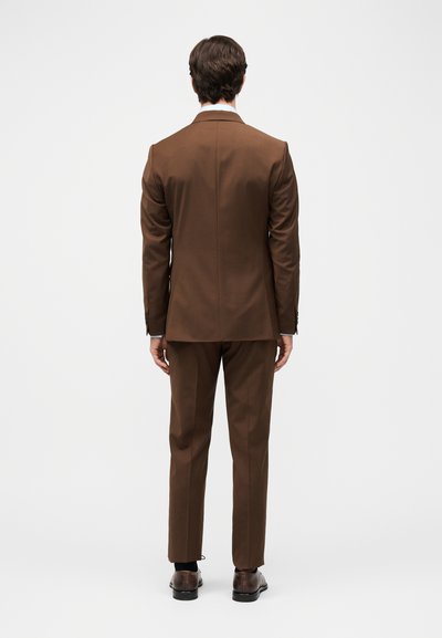 Άνδρας όρθιος με την πλάτη, φοράει καφέ κοστούμι με παντελόνι που ταιριάζει και σκούρα καφέ επίσημα παπούτσια σε λευκό φόντο.