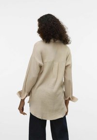 Beige, textured blouse with a relaxed fit, long sleeves, and a back pleat detail. Worn with dark blue pants, showing a back view.