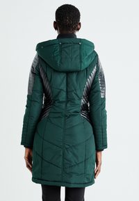 Garšs zaļš pūkains mētelis ar kapuci, kam ir melni kvadrātveida paneļi sānos un piedurknēs, pieguļošs siluets un ribotas aproces.