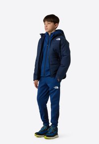 Laivastonsininen toppatakki hupulla, päällä samansävyinen sininen vetoketjullinen paita ja collegehousut. Mustat ja vihreät vaelluskengät, joissa on teksturoidut pinnat ja nauhat.