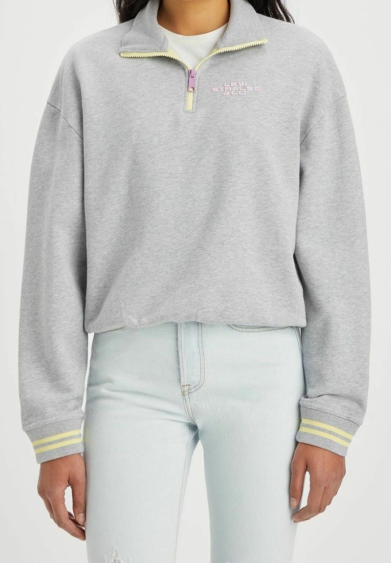 Sudadera gris con cuello de media cremallera, cremallera amarilla y logo bordado en rosa. Presenta puños a rayas y un ajuste relajado.