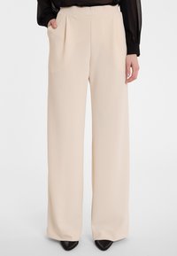 Beige wijde broek met voorplooien, zachte stof en zijzakken. Glad textuur, hoge taille, gecombineerd met zwarte puntige schoenen.