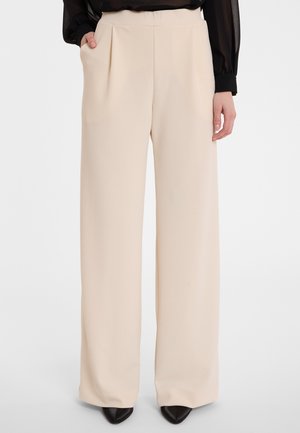 Beige weit geschnittene Hose mit vorderen Falten, aus weichem Stoff und mit seitlichen Taschen. Glatte Textur, hohe Taille, kombiniert mit spitzen schwarzen Schuhen.
