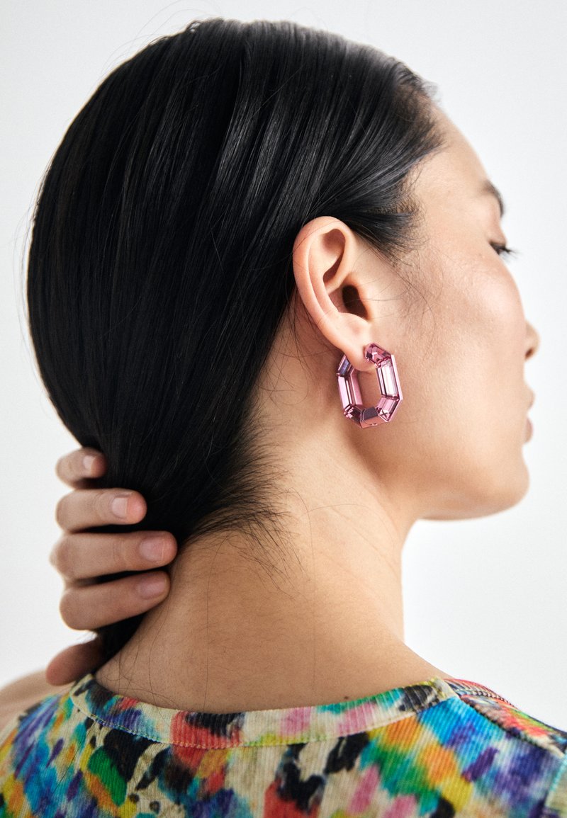 Large pink geometric earrings made of acrylic, featuring an octagonal shape with faceted edges, worn by a person with long dark hair.