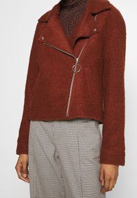 Giacca di colore ruggine, con texture, colletto a punta, zip asimmetrica e tasche laterali; abbinata a pantaloni a quadri.