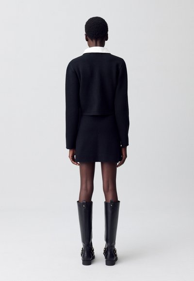 Pull noir court à manches longues, associé à une jupe mini noire et de longues bottes noires brillantes avec des accents en métal doré.