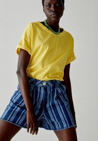 T-shirt di cotone gialla con maniche arrotolate, abbinata a pantaloni corti in denim a strisce blu e bianche con chiusura a bottone e tasche frontali.