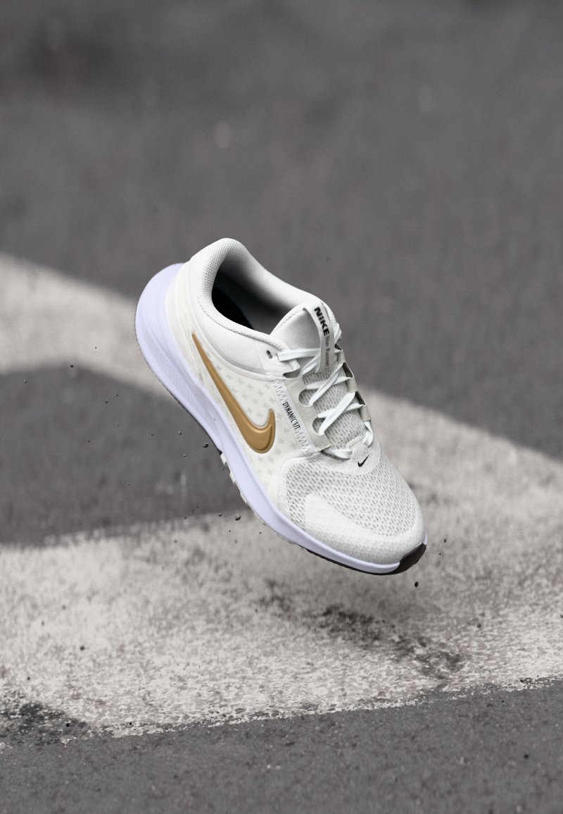 Baskets de sport gris clair avec tige en mesh et swoosh doré, dotées d'une semelle blanche et d'accents texturés, positionnées au-dessus d'un passage piéton.