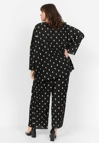 Schwarzes und beigefarbenes gepunktetes Outfit aus plissiertem Stoff, bestehend aus einer lockeren Bluse und weit geschnittenen Hosen. Betont mit einer lässigen Silhouette.