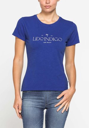Donna che indossa una maglietta blu aderente con testo bianco "Lido Indigo Just Relax" e jeans blu, in piedi con una mano vicino all'anca.