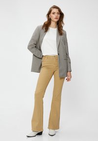 Grau karierter Blazer über einem weißen T-Shirt, kombiniert mit beigen, ausgestellten Hosen und weißen Knöchelstiefeln. Glatte Textur mit strukturierten Designelementen.