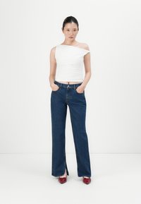 Hvit, avskåret topp uten skuldre, kombinert med høyt liv og rette ben i mørkeblå jeans. Modellen står med hendene i lommene og har på seg burgunder sko.