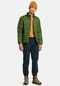 Groene pufferjack met ritssluiting, gedragen over een mosterdkleurige trui, gecombineerd met marineblauwe broeken en bruine wandelschoenen. Muts met logodetail.