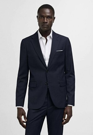 Navy blue tailored suit jacket with a notched lapel, two buttons, and side pockets. Worn over a white dress shirt with a pocket square.