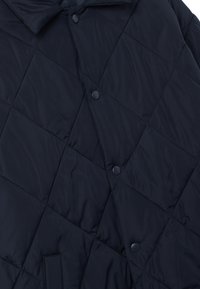 Giacca trapuntata blu navy con un motivo a diamante, bottoni a pressione e colletto alto. Il tessuto sembra leggero e presenta una texture liscia.