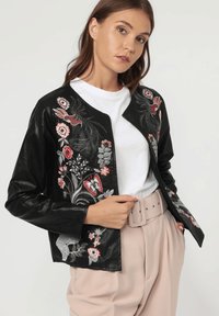 Chaqueta de cuero negra con bordados florales coloridos. Presenta un cuello redondo y mangas largas. Combinada con una camisa blanca y pantalones beige.