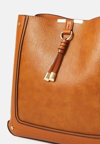 Brun lædertaske med struktureret finish, der har guldfarvet hardware, en sikret kvastdetalje og en kontrasterende sort kant.