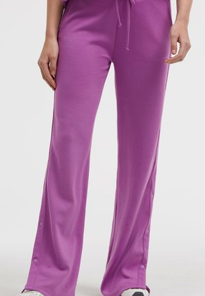 Person trägt lockere violette Hose mit Druckknöpfen entlang der Außenseiten, kombiniert mit weißen Turnschuhen, steht vor einem schlichten Hintergrund.