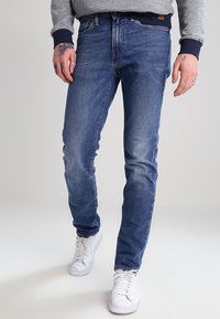 Slim-fit blå denimjeans med lätt slitage, fem fickor och kontrasterande sömmar, kombinerat med vita sneakers och en grå tröja.