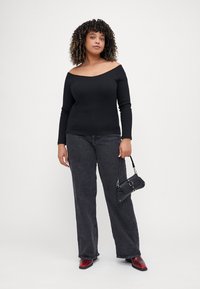 Top negro de canalé con hombros al aire y mangas largas, combinado con unos jeans oscuros sueltos grises y botas rojas de tacón, sosteniendo un pequeño bolso negro.