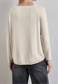 Beżowy polarowy pulower w jasnym odcieniu beżu, z długimi rękawami raglanowymi i zaokrąglonym brzegiem, charakteryzujący się miękką teksturą i minimalnymi przeszyciami.