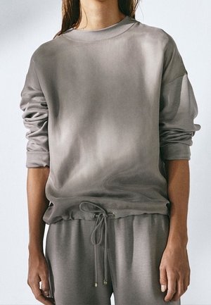 Nainen pukeutuneena löysään harmaaseen pitkähihnaiseen collegepaitaan ja harmaisiin collegehousuihin, joissa on kiristysnaru vyötäröllä, seisoo yksiväristä taustaa vasten.